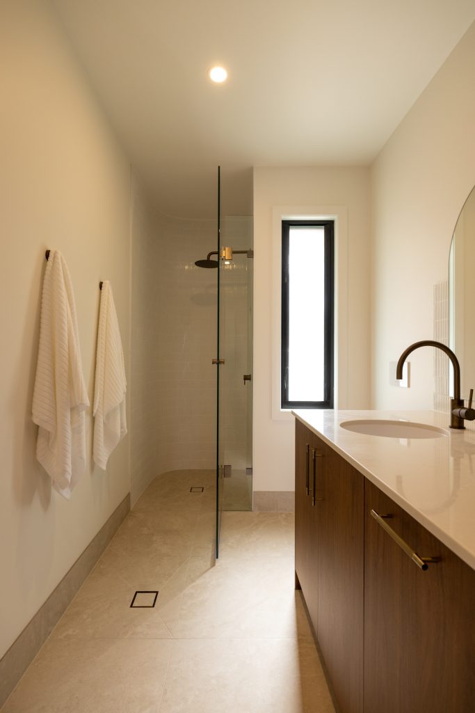 A key part of getting a combined space right is ensuring the bathroom experience is not compromised. Here, the basin, mirror and tapware have been treated with the same level of attention you would expect in a main bathroom. The curved mirror softens the joinery lines, while the textured splashback adds detail without overwhelming the space. The walk in shower is generous and clean, with minimal framing and a layout that feels open and easy to use. Nothing feels like an add on. The laundry components are integrated so seamlessly that the space reads as a bathroom first, with the laundry quietly built into it.