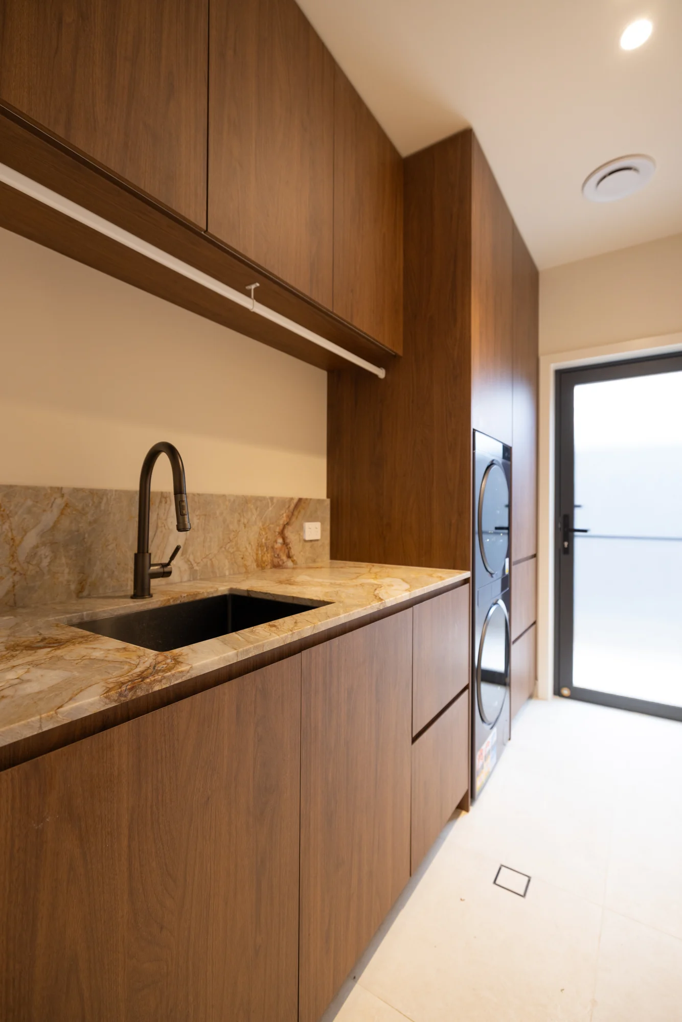 Solara House laundry with warm timber-tone cabinetry, overhead cupboards, sink and stacked appliances